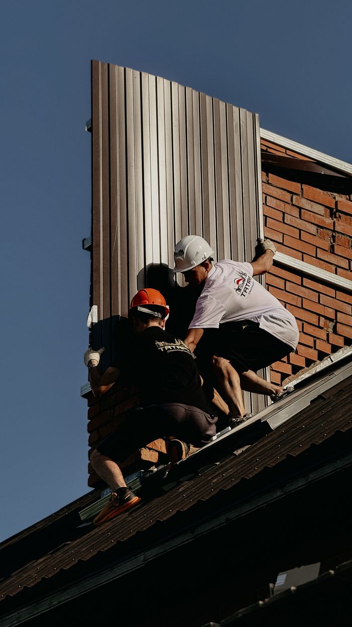 Two construction workers are installing metal panels on a brick building roof.
